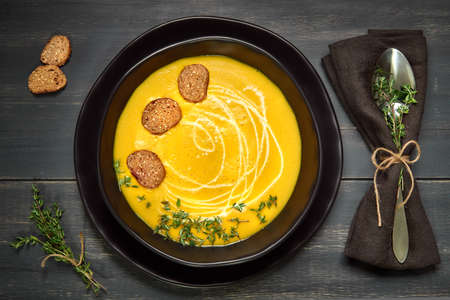 Pumpkin creme soup in ceramic bowl served with wholegrain croutons, thyme leaves and cream on dark wood, top viewの写真素材