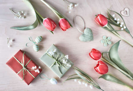 Overhead view of light wooden table with springtime decorations, wrapped gifts, white flowers, red tulips and and Easter eggs. Spring or Easter background concept. の写真素材