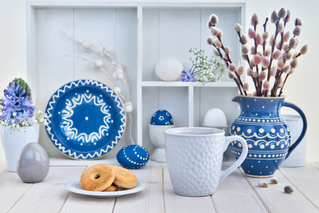 Cup of coffee and cookies on white table with bunch of pussy-willow in blue ceramic jar and matching springtime decorationsの写真素材