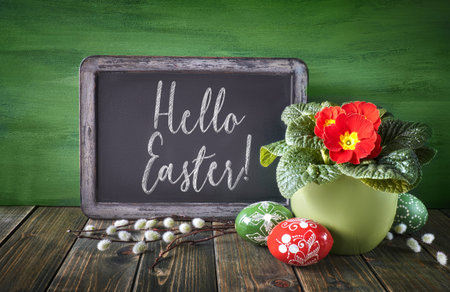 Easter basket with painted Easter eggs and red primrose pot flower on rustic wooden background, text on blackboardの写真素材
