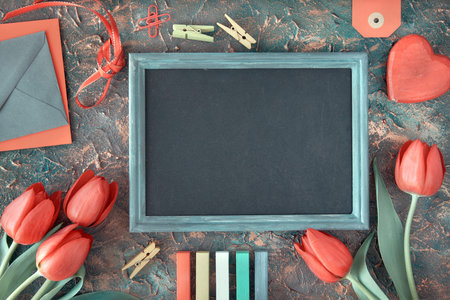 Spring holiday background: blackboard framed with red tulips, wooden heart, color chalks and...