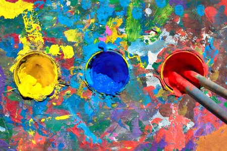 Gouache paint table with paint smears and cups with yellow, blue and red paints. Top view, two...