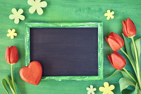 Spring holiday background: blackboard framed with red tulips, wooden heart and flowers on green...