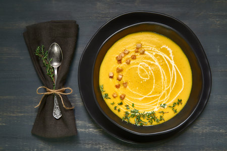 Pumpkin creme soup in black ceramic bowl served with wholegrain croutons, thyme leaves and cream on...