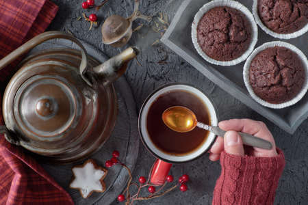 Female hand mixes cup of tea with a spoon on a cold morning in Fall. Vintage kettle, tea mesh, cookies and chocolate muffin, Autumn leaves and red and berries on dark background, flat layout.の写真素材