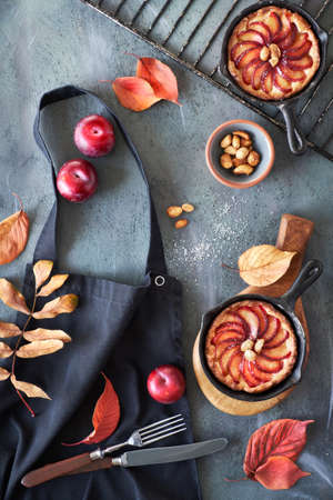 Red plums and plum tarts in cast iron skillets on a black cooking apron thrown on gray textured background, Autumn baking concept, flat lay compositionの写真素材