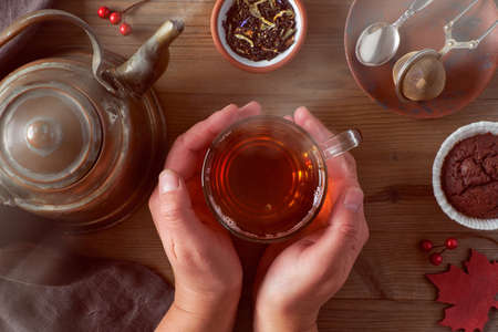 Female hand holding a warm cup of tea on a cold morning in Fall. Vintage kettle, spoon, tea mesh, chocolate muffin, Autumn leaves and red and berries on dark background, flat layout.の写真素材