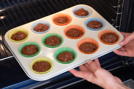 Female hands setting baking tray with raw chocolate dough in colorful paper cups into oven. Concept...