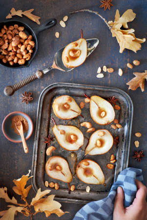 Flat lay with a tray of baked pears with caramelized nuts on dark wooden background with Autumn leaves, sugar and honeyの写真素材