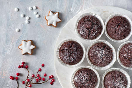 Flat lay with red berry decoration on white rustic background and chocolate muffins in white paper cups with sugar icing on white marmor boardの写真素材