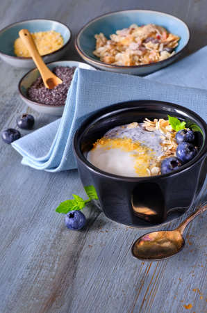 Yogurt with muesli, blueberries, poppy seeds, mint and crushed cornflakes served in black shiny bowl on rustic wooden tableの写真素材