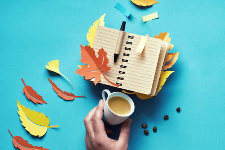 Creative top view flat lay on the topic of Autumn planning and to-do lists. Hand holding offee cup and notebook with colorful bookmarks as Autumn leaves on blue paper background.の写真素材
