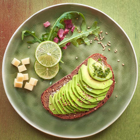 Flat lay of avocado sandwich and green salad with ham cubes on brown-green textured background, top view, square compositionの写真素材