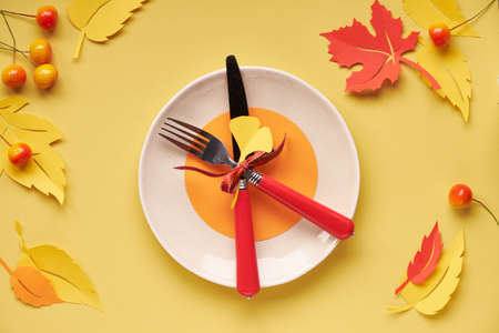 Table setup for Autumn celebration, Thanksgiving or birthday. Ceramic plate with fork and knife on yellow paper with paper Autumn leaves and berriesの写真素材