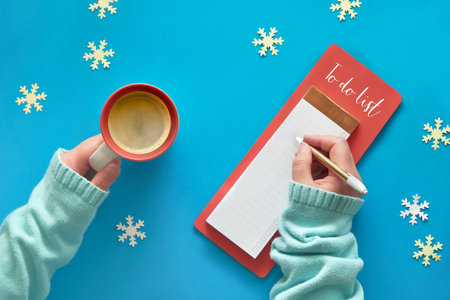 Christmas do list and woman's hands in mint color sweater with cup of coffee on light blue pastel...