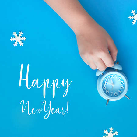 Child's hand holding blue alarm clock showing five to twelve with paper snowflakes. New Year's night, midnight, flat lay concept image, top view on pastel blue paper background.の写真素材