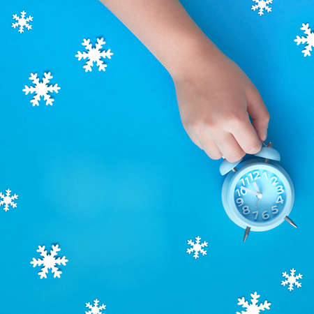 Child's hand holding blue alarm clock showing five to twelve. New Year's night, midnight. Flat lay concept image, top view on pastel blue paper background with white snowflakes, square composition with copy-space.の写真素材