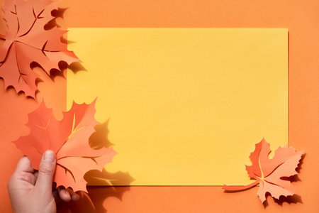 Hand holding range maple leaf on paper background with shadows and copyspace in paper frame. Creative handmade paper craft, flat lay, Autumn paper background.の写真素材