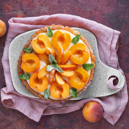 Top view of apricot tart with mint leaf on ceramic board with linen towel on dark red textured backgroundの写真素材