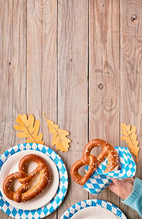 Oktoberfest, female hand in blue sweater holding pretzel. Flat lay on rustic wooden table with blue and white checkered disposable paper plates, orange Autumn leaves and copy-space.の写真素材