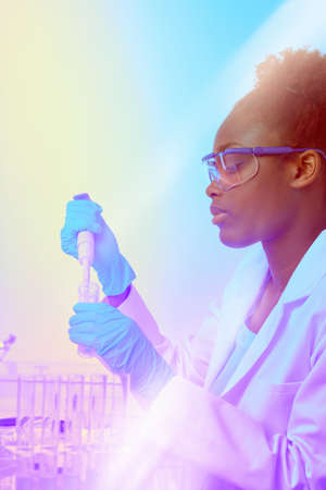 African scientist or graduate student in lab coat and protective wear works with automatic pipette in modernl laboratory. Futuristic neon glow, abstract light pattern in purple, blue and yellow, text space.の写真素材