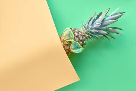 Funny pineapple in heart-shaped glasses on geometric diagonal paper background in lime green and yellow colors with copy-spaceの写真素材