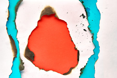 Burnt hole in white and turquoise paper with burned edges, flat lay on vibrant orange paper with...