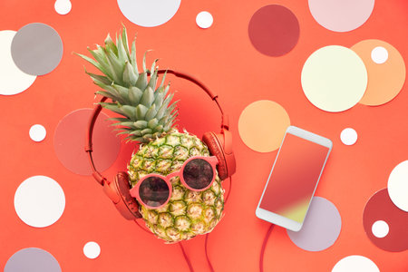 Funny pineapple in sunglasses and earphones painted blue. Flat lay on red paper background with...