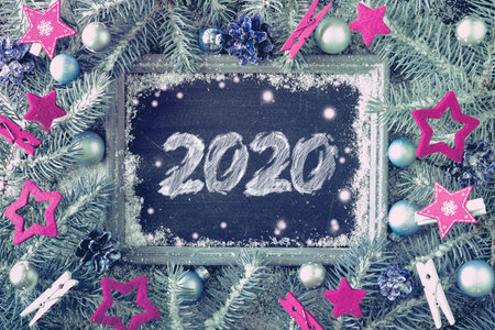 Happy New Year 2020! Decorated fir twigs around chalk board on rustic wood with snow. Top view with...