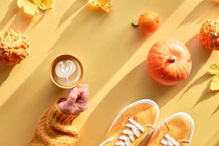 Yellow Autumn paper flat lay with hand with cup of pumpkin spice latte, orange pumpkins, maple...
