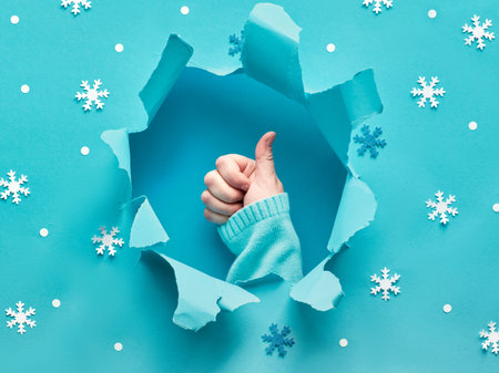 Blue paper flat lay with snowflakes and ripped hole in the middle. Female hand showing OK sign. All...