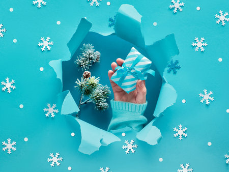 Blue paper flat lay with snowflakes and ripped hole in the middle. Female hand in the hole holding...