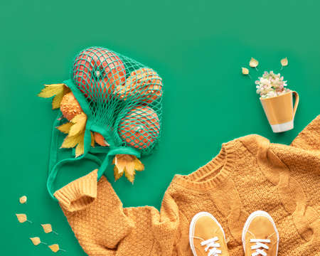 Autumn flat lay on green paper background with copy-space. Orange pumpkins in mesh bag. Mug with flowers. A pair of canvas sneakers on orange sweater holding the net bag.の写真素材