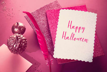 Creative purple and pink Halloween background with levitating pink pin quill, stack of glittering paper and decorative pumpkins painted metallic pink. Text "Happy Halloween" on silver paper.の写真素材