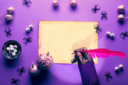 Creative Halloween background in purple, pink and black. Witch in black mesh gloves writing on parchment with quill. Flat lay with spiders, pink pumpkins and chocolate eyes, space for your text.の写真素材