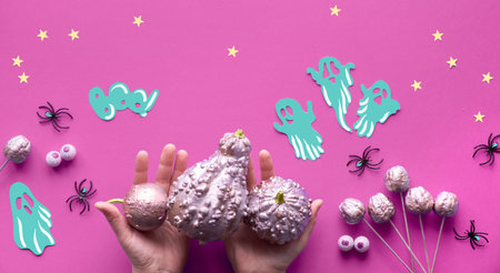 Creative Halloween flat lay on purple paper background with paper ghosts, stars and chocolate eyes. Hands in black mesh gloves holding gilded pink pumpkins.の写真素材