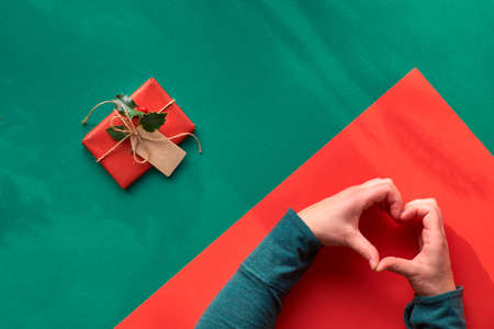 Creative diagonal geometric flat lay on green and red paper with long floral shadows . Wrapped gift box decorated with holly. Hands showing heart sign. Eco-friendly gift ideas for Xmas.の写真素材