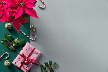 Christmas flat lay on split silver grey and green paper background. Vibrant red poinsettia plant, candy canes, holly twigs, dry lime fruit and pink wrapped Xmas gift with trinketの写真素材