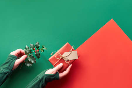Geometric flat lay on green and red paper, copy-space. Female hands holding wrapped gift box and twig of holly with red berries. Eco friendly gift ideas for zero waste Xmas.の写真素材