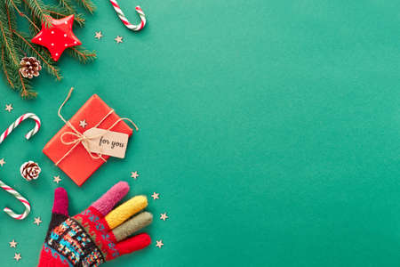 Creative Christmas flat lay background in green and red on green paper with copy-space. Hand in ornate wool glove, candy canes, wrapped gift with cardboard tag, stars and fir twigs.の写真素材