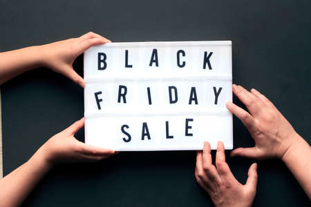 Lightbox text "Black Friday Sale" on black paper background. Creative flat lay with two pairs of hands, child and adult, holding and touching the light box.の写真素材