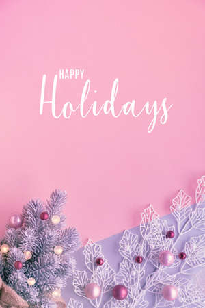 White winter twig with shiny geometric leaves and artificial Xmas tree decorated with lights and glass baubles. Christmas flat lay on two color paper, pink and silver. Xmas text "Happy Holidays".の写真素材