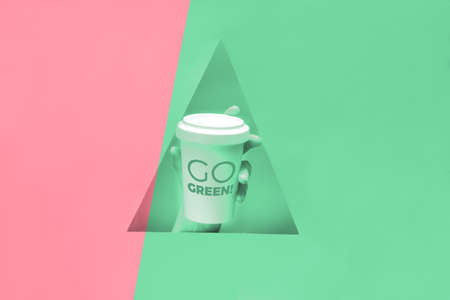 Reusable coffee travel mug, bamboo cup with text "Go green" in hand showing through triangular paper hole. Split pink and green two color monochrome background, toned image.の写真素材