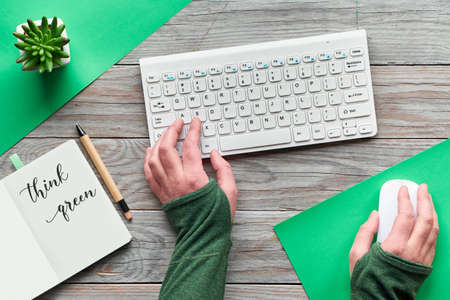 Think green, zero waste creative flat lay. Top view with hands typing on keyboard and holding computer mouse. Open notebook with motivation text, minimal wooden workspace with succulent plant on wood.の写真素材