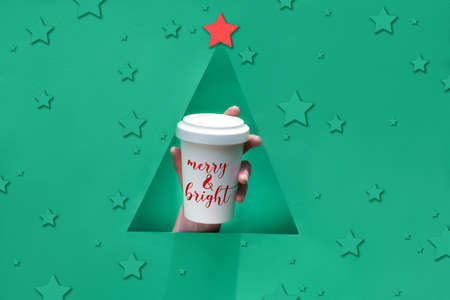 Stylish reusable eco coffee travel mug, bamboo cup with lid in hand through triangular paper hole in shape of Christmas tree. Green paper background, keep cup with text "Merry and bright".の写真素材