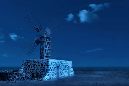 Classic blue monochrome toned image of traditional windmill at night with moonlight by the calm Atlantic ocean. Fuerteventura, Canary islands, Spain. Calm night with clear sky and clouds.の写真素材