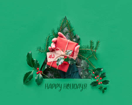 Hand showing wrapped gift in triangular paper hole in a shape of Christmas tree with red paper star. Natural holly and fir twigs. Creative minimalist Xmas flat lay in trendy neo mint green and red.の写真素材