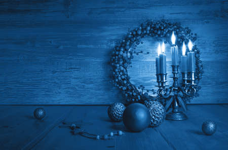 Trendy blue monochrome Xmas composition with candles in candelabrum, berry wreath and classic vintage glass baubles. Vintage Christmas decorations on wood with copy-space.の写真素材