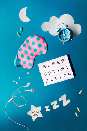 Healthy night sleep creative concept with text "Sleep optimization" on lightboard. Flying or levitating objects: sleeping mask, alarm clock, earphones, earplugs and pills. Paper moon and star in hand.の写真素材