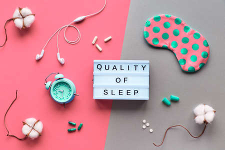 Text "Quality of sleep", creative concept. Sleeping mask in polka dots, small alarm clock, earphones and earplugs. Pills and capsules with sleep remedy. Flat lay, top view, split two tone backgroundの写真素材